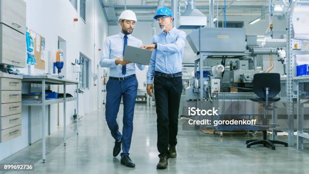 Back View Of The Head Of The Project Holds Laptop And Discussing Product Details With Chief Engineer While They Walk Through Modern Factory Stock Photo - Download Image Now