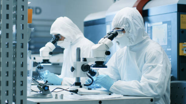 two engineers/ scientists/ technicians in sterile cleanroom suits use microscopes for component adjustment and research. they work in an electronic components manufacturing factory. - smiling research science and technology clothing imagens e fotografias de stock