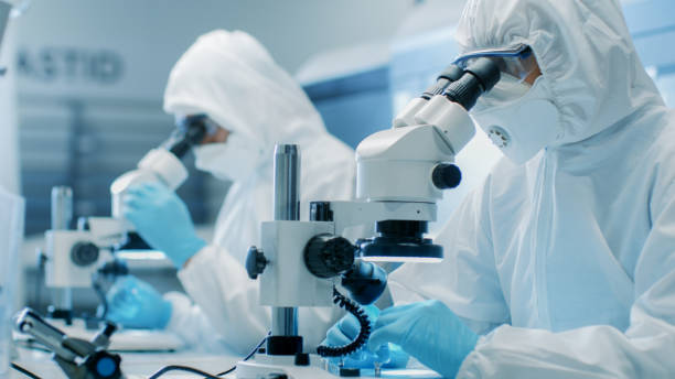 two engineers/ scientists/ technicians in sterile cleanroom suits use microscopes for component adjustment and research. they work in an electronic components manufacturing factory. - smiling research science and technology clothing imagens e fotografias de stock
