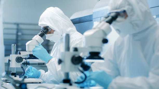 two engineers/ scientists/ technicians in sterile cleanroom suits  control manufacturing machinery work and use microscopes for component adjustment and research. they work in an electronic components manufacturing factory. - smiling research science and technology clothing imagens e fotografias de stock