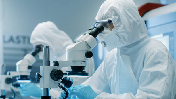 istock Two Engineers/ Scientists/ Technicians in Sterile Cleanroom Suits  Control Manufacturing Machinery Work and Use Microscopes for Component Adjustment and Research. They Work in an Electronic Components Manufacturing Factory. 899692984