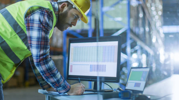 istock Male Inspector Fills up Spreadsheets on His Personal Computer. He's in Big Warehouse with Rows of Pallet Racks. 899693144
