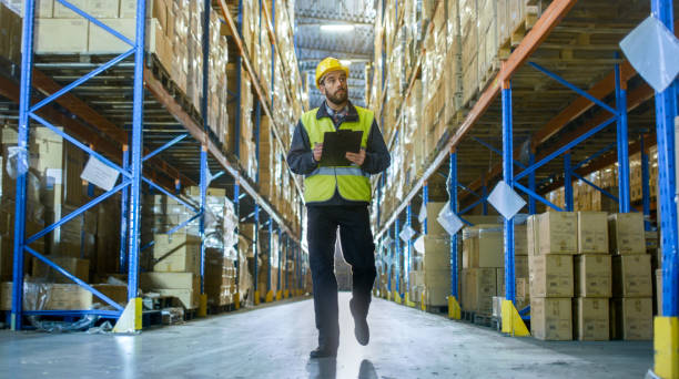 istock Overseer Wearing Hard Hat with Tablet Computer Counts Merchandise in Warehouse. He Walks Through Rows of Storage Racks with Merchandise. 899693180