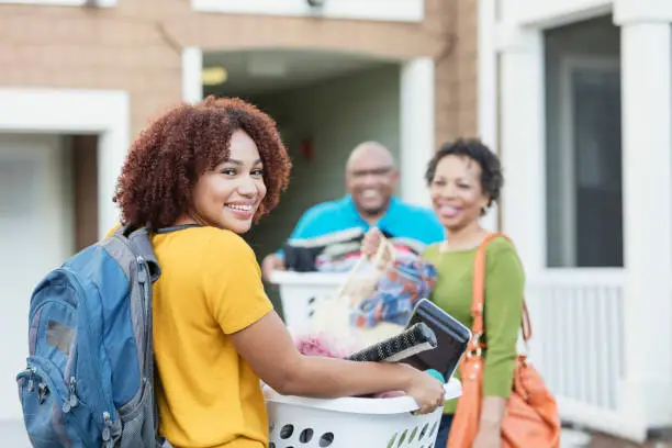 Mature African-American parents helping their daughter relocate, perhaps into an apartment or college dorm.  The young woman is in the foreground smiling at the camera, carrying a backpack and basket filled with her belongings.