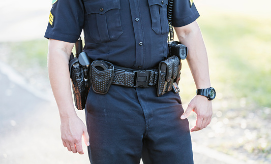 Cropped view of a police officer's waist. He is wearing his equipment belt with gun holster, handcuffs, radio and other gear. He is an Hispanic mid adult man in his 30s standing outdoors.