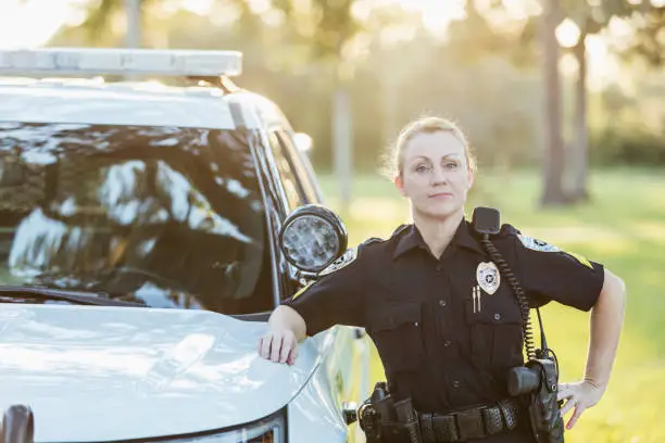 A policewoman standing beside her squad car, leaning against it, looking at the camera with her hand on her hip. She is a mature woman in her 40s.