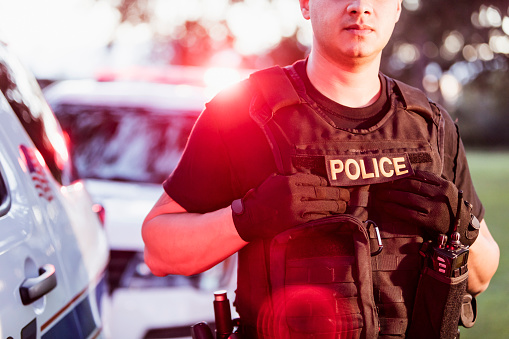 Cropped view of an Hispanic police officer wearing a bulletproof vest, standing beside two police cars. He is a mid adult man in his 30s.