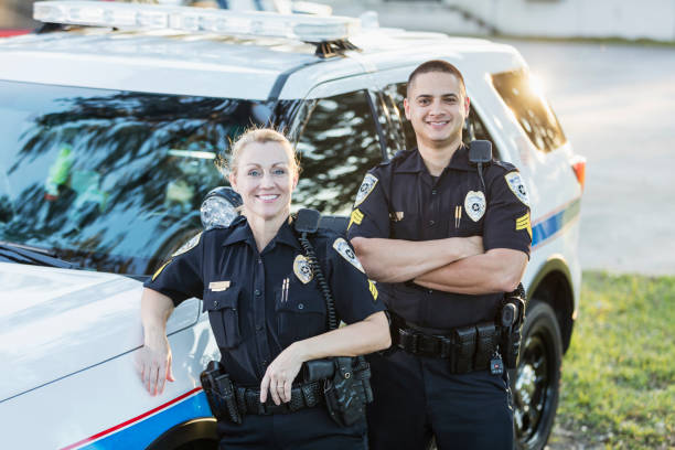 Policewoman and partner next to squad car A policewoman standing beside her squad car, leaning against it, looking at the camera. She is a mature woman in her 40s. Her partner, an Hispanic man in his 30s, is standing behind her, with his arms crossed. police force smiling officer stock pictures, royalty-free photos & images