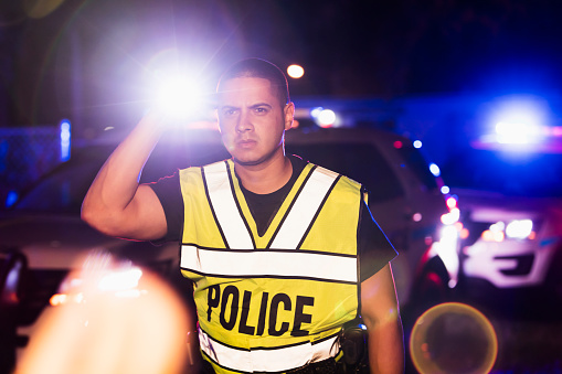 An Hispanic police officer wearing a reflective vest, standing in front of police cars with emergency lights flashing, looking away with a serious expression, holding a flashlight. He is a mid adult man in his 30s.