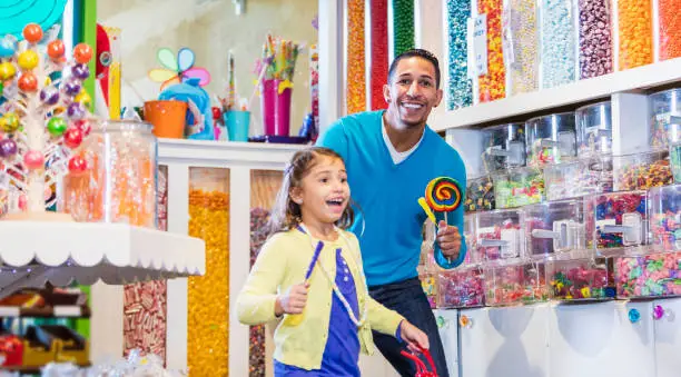 A little 7 year old girl smiling with her father, standing in a candy store, surrounded by jars, containers and displays of colorful candies. They are mixed race Hispanic and Caucasian.