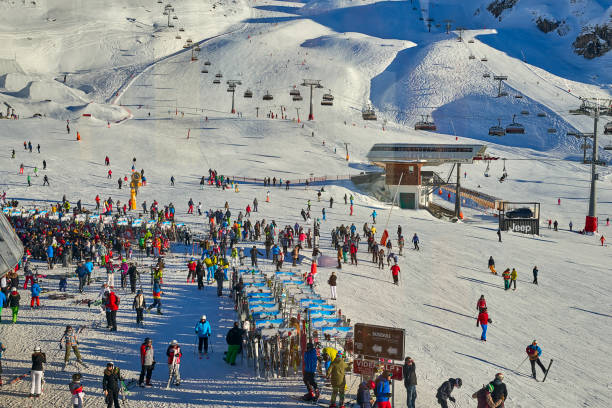 Ischgl, Austria - December 25, 2017: Ski base in Tyrol Alps in sunny December day. Chairlifts and cable cars are full of people, crowd is in front of entrance. Foreground shows rack for skis, where skiers can leave skis and relax in cafe.
