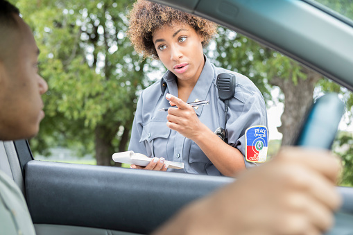 A young female police officer points at a male driver as she discusses the traffic laws that he has violated.  He is unrecognizable and she holds a note pad and pen.  This photo is taken from the point of view of a front seat passenger.