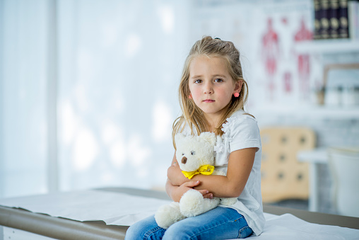 A young Caucasian girl is indoors in a medical clinic. She is holding a teddy bear in her arms. She is sitting and looking worried about her checkup.