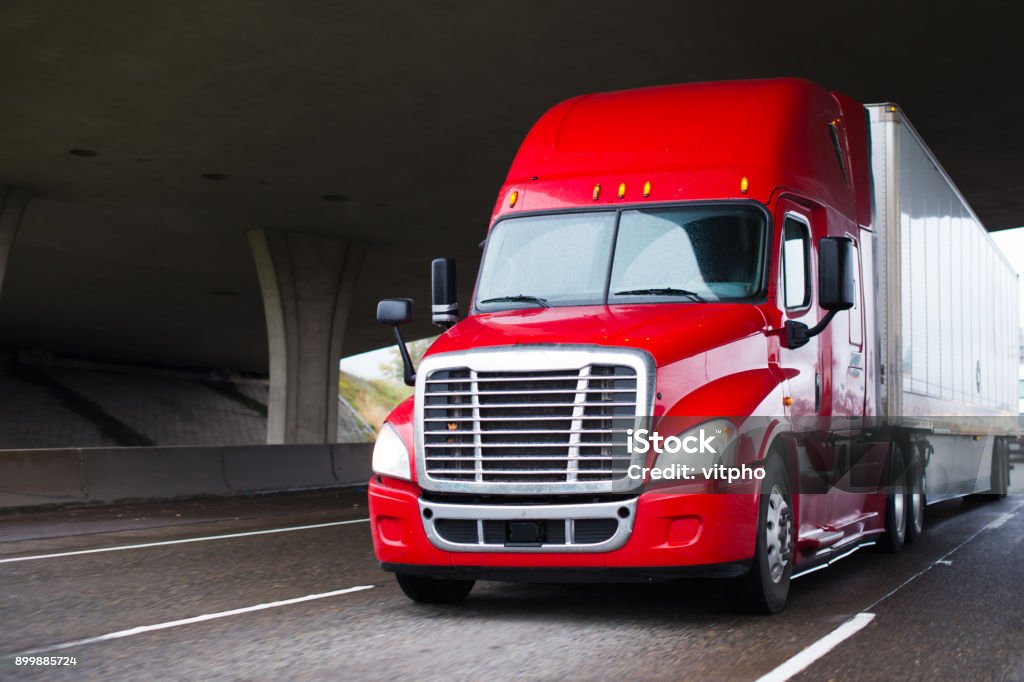 Bright red modern big rig semi truck with dry van trailer running under concrete bridge A modern big rig semi truck for long haulage with a high cabin for improving aerodynamic characteristics moves under the bridge across a multi-lane highway transporting a dry van semi trailer with commercial cargo to the place of delivery Truck Stock Photo