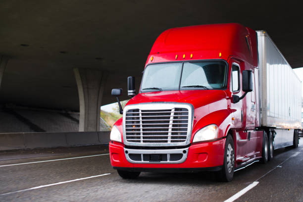 Bright red modern big rig semi truck with dry van trailer running under concrete bridge A modern big rig semi truck for long haulage with a high cabin for improving aerodynamic characteristics moves under the bridge across a multi-lane highway transporting a dry van semi trailer with commercial cargo to the place of delivery aerodynamic photos stock pictures, royalty-free photos & images