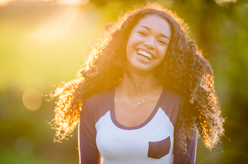 A teenage girl of African descent is outdoors. She is smiling at the camera while the sun shines behind her.