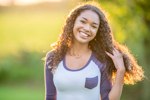 A teenage girl of African descent is outdoors. She is smiling at the camera and touching her hair while the sun shines behind her.