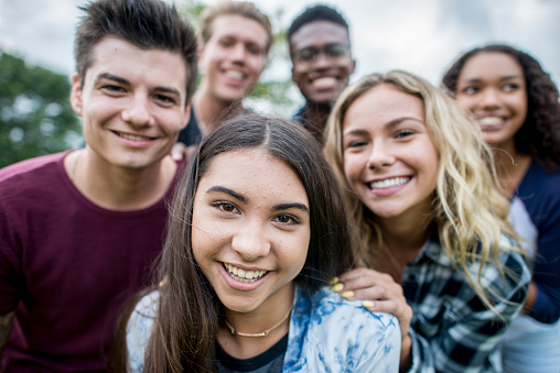 A multi-ethnic group of high school students are outdoors on a summer day. One girl is taking a selfie of the whole group. Everyone is smiling.