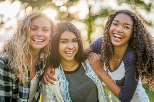 Three teenage girls are outdoors on a summer day. They are all smiling at the camera while the sun sets through the trees behind them.