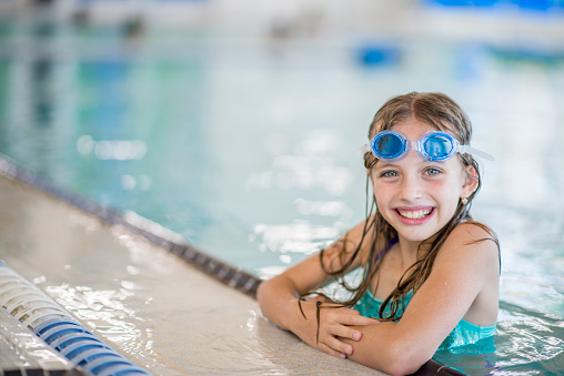 A Caucasian girl is swimming indoors at a fitness center. She is smiling at the camera while at the edge of the swimming pool. She is wearing goggles.