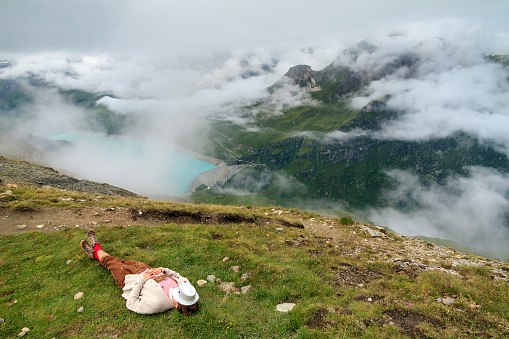 A young man in traditional clothing with lederhosen and checkered shirt takes a nap on the grass in summer in the alps near Grimentz, Switzerland, with turquoise lake Lac de Moiry in the background