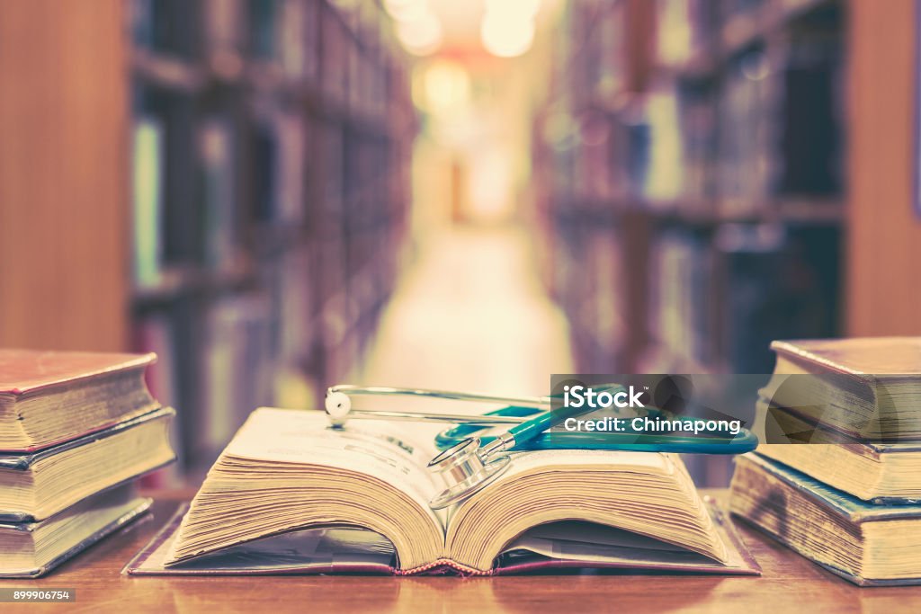 Medical library concept with stethoscope on book Old book in library with stethoscope on open textbook, stack piles of texts on reading desk, and aisle of bookshelves in  study class room background for medical school education learning concept Healthcare And Medicine Stock Photo