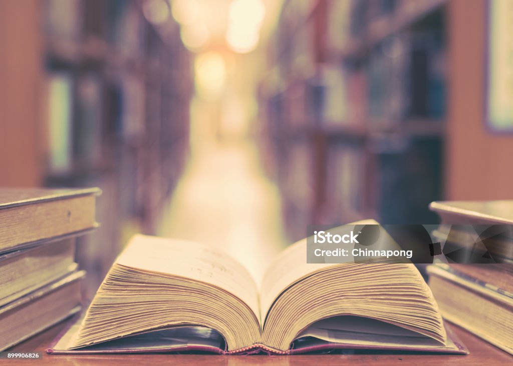 Education concept with old book in library Old book in library with open textbook, stack piles of literature text archive on reading desk, and blur aisle of bookshelves in school study class room background for education learning concept Philosophy Stock Photo