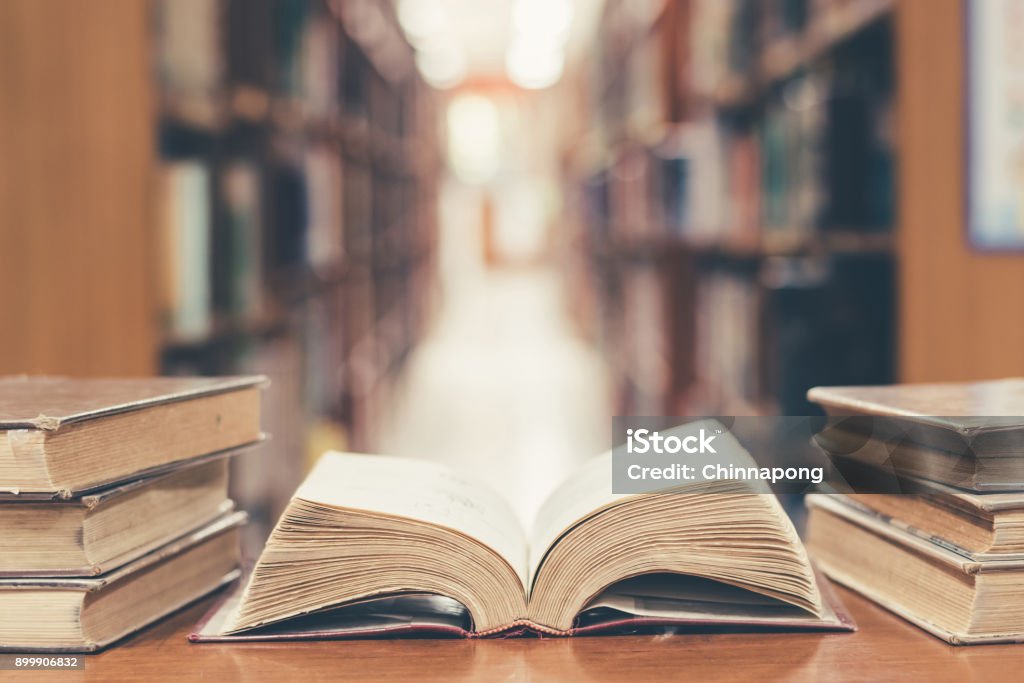 Education concept with old book in library Old book in library with open textbook, stack piles of literature text archive on reading desk, and blur aisle of bookshelves in school study class room background for education learning concept Book Stock Photo