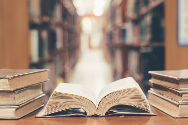 Education concept with old book in library Old book in library with open textbook, stack piles of literature text archive on reading desk, and blur aisle of bookshelves in school study class room background for education learning concept human interest stock pictures, royalty-free photos & images