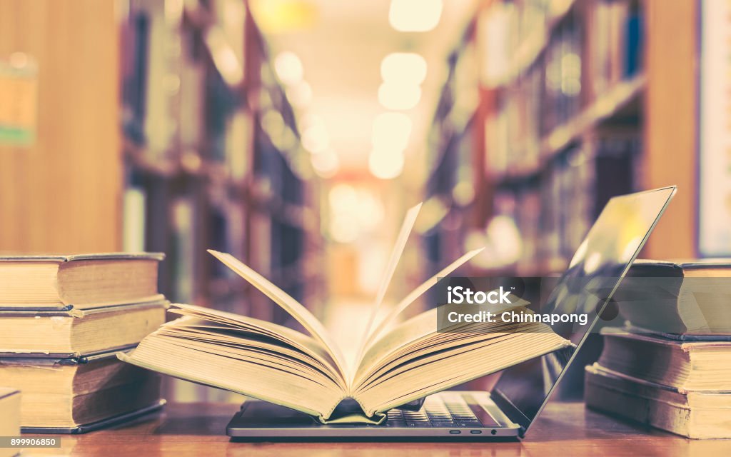 Book and computer technology in library Education e-learning class and e-book digital technology concept with computer notebook and opening book flipping on classroom desk in school library among old stacks of textbook archive collection Book Stock Photo