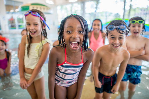 A multi-ethnic group of kids are posing together by the edge of a swimming pool. Some kids are standing in the water. They are all smiling at the camera.