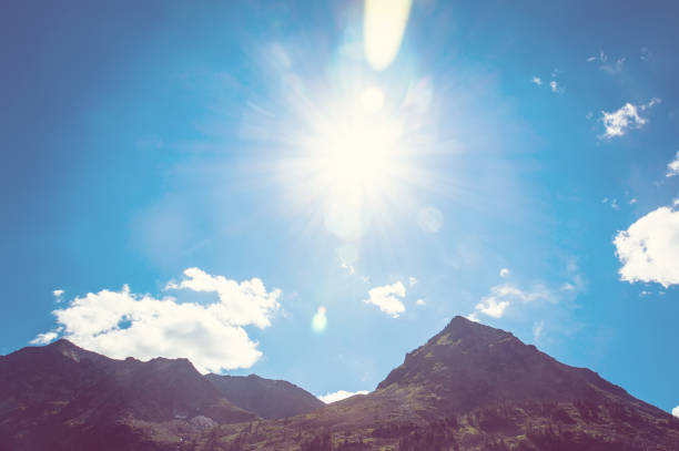 top of mountains in blue sky. The sun shines brightly over the top of a high mountain on the background blue beautiful sky with white clouds