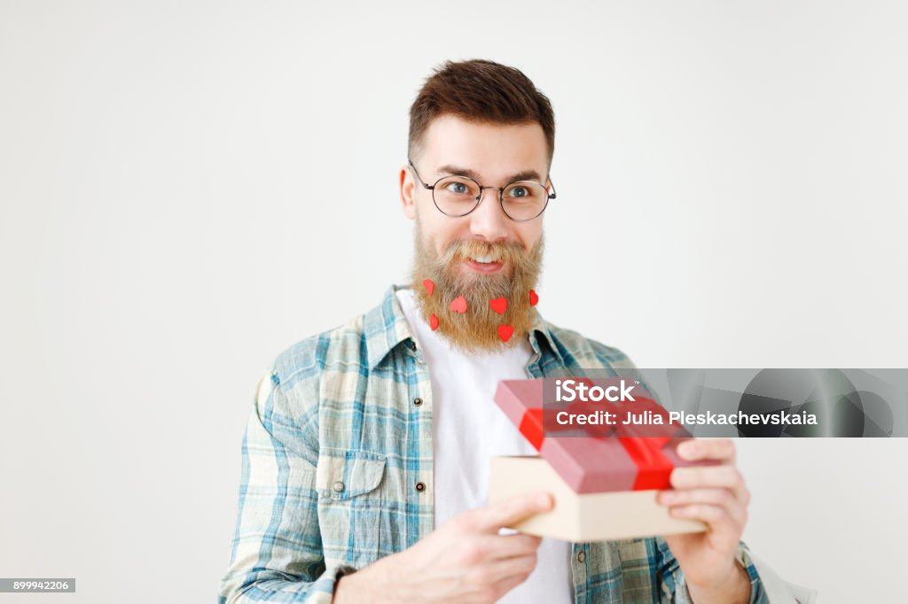 Fashionable bearded hipster guy in eyewear, opens wrapped present, recieves surprise from wife, being in good festive mood, isolated over white background. People, surprisment and presents concept Fashionable bearded hipster guy in eyewear, opens wrapped present, recieves surprise from wife, being in good festive mood, isolated over white background. People, surprisment and presents concept. Adult Stock Photo