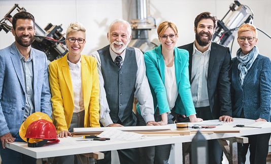 Closeup  front view of mixed age people of industrial design department at a factory. They are gathered at a desk in large production facility and discussing certain blueprints and documents. At the moment they are looking at the camera. There are three men and three women.