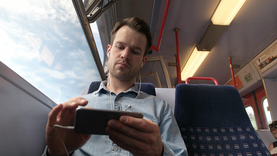 A good looking young man, sitting on a train, using his smartphone to watch a movie or browse the internet with headphones plugged in.