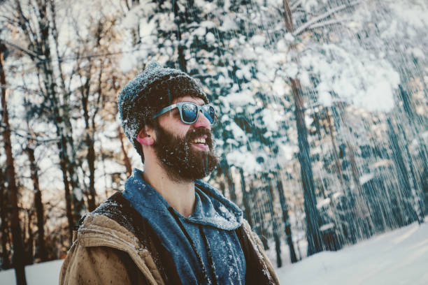One hipster beard man is standing in deep snow. This man is half covered by snow, so he had a funny day in the snow. He is on winter journey and have amazing adventure time because it's snow all around him. Behind him is pine tree woodland also covered by snow. It's cold outside so he is wearing a lot of warmth clothes, hat, gloves and sunglasses.