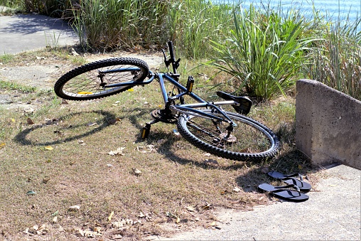 Bicycle thrown on ground beside pair of flip flops. In background is bush and water.