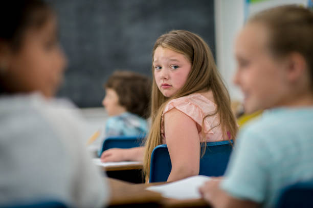 Being Bullied An elementary school girl is being bullied in her classroom. Two other girls are whispering about her. The first girl is looking at them sadly. sad 15 years old girl stock pictures, royalty-free photos & images