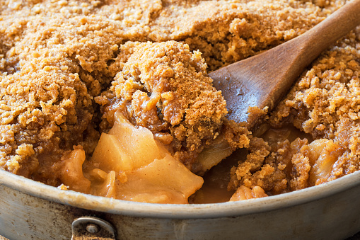 A close up macro shot of a wooden baking spoon scooping out a piece of Apple Crisp or Apple Crumble from a metal baking pan. This dessert is made from spiced apples with a brown sugar crumble topping.