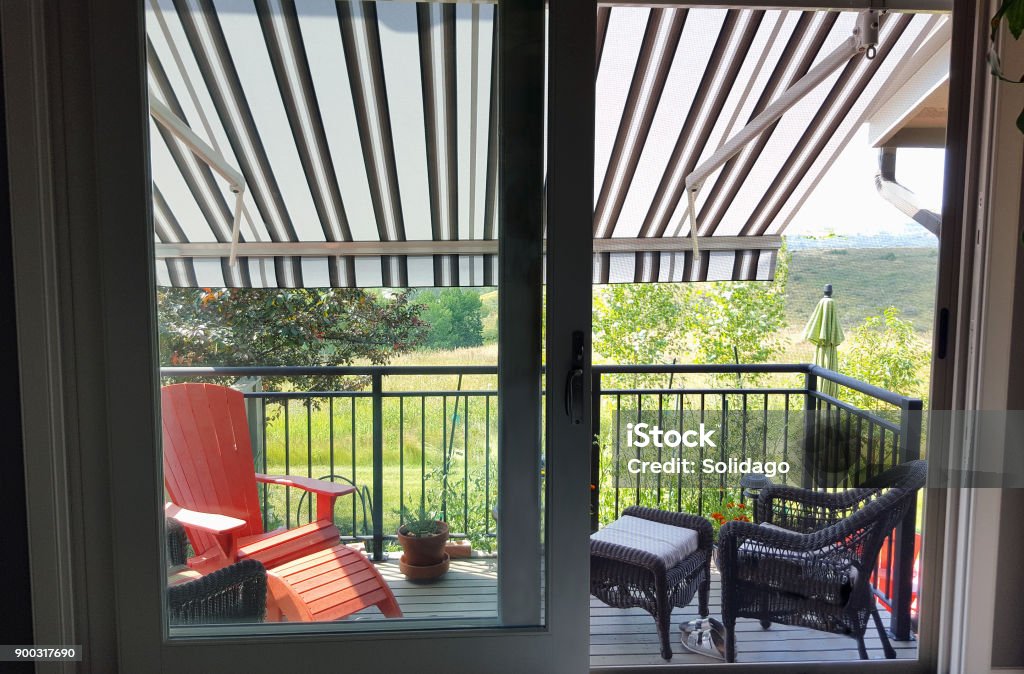 Looking Out To A Cool Awning Shaded Deck Property Released. Inside looking out to residential deck with awning.  Wicker furniture and a red Adirondack chair on deck. Garden with trees below. Awning Stock Photo