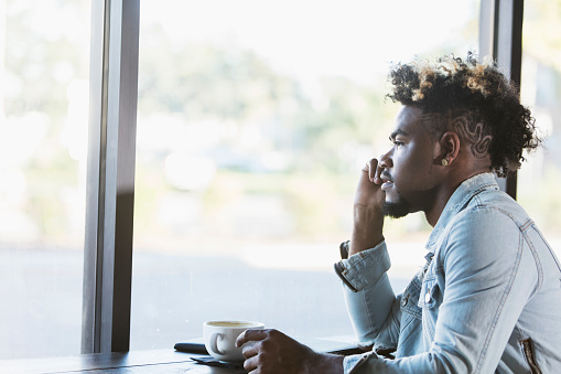 A young mixed race man in his 20s sitting by the window in a coffee shop, thinking. He has an earring, a beard and an afro with a few streaks of dyed blond hair, and a shaved pattern on his scalp.