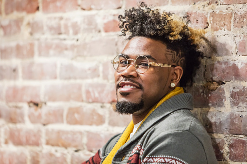Close-up of a young, mixed race African American and Caucasian man wearing stylish casual clothing, eyeglasses and an earring leaning against a brick wall. His hair is curly, dyed with streaks of light brown. He is smiling confidently at the camera.