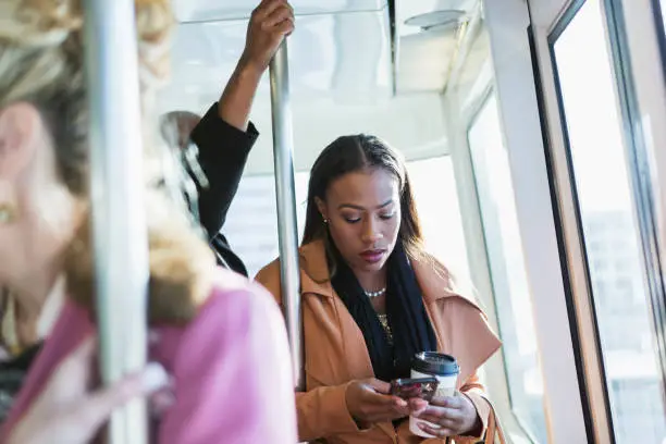 An African-American woman in her 30s riding the train during rush hour, holding a cup of coffee in a disposable cup, texting on her mobile phone, people out of focus around her. She has a serious expression on her face.