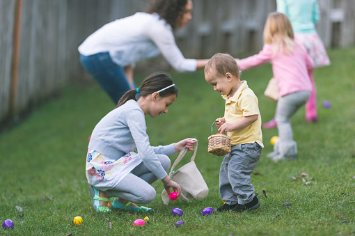 Children participate in Easter games in a residential, fenced-in backyard. A kind older sister helps her younger brother find some plastic colored eggs laying in the grass.