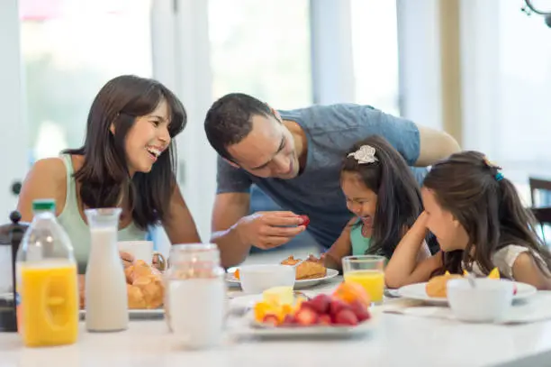 Two ethnic parents try to survive breakfast with their two mischieous daughters. There is a spread of fresh fruit and croissants on the table. Dad is leaning over the younger girl and trying to give her a strawberry...and she is refusing. All are smiling or laughing.