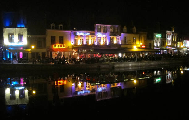 Amiens, France - April 8, 2017: People sit at cafe tables beside the river, surrounded by colorful night lights of shops and restaurants. Amiens, located in north-west France, is the capital city of the Somme department and is situated on the banks of the Somme River.