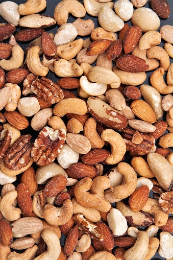 Lightly salted mixed nuts spread out on a dark background. Includes cashews, almonds, pecans, brazil nuts, hazelnuts, macadamia nuts. Background image.