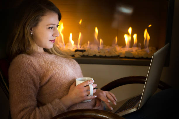 istock Young woman at a fireplace, using laptop and having coffee. 900479588