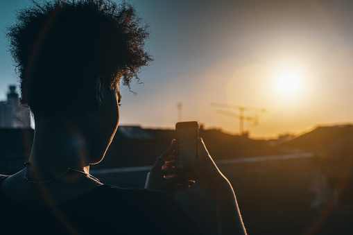 Rear view of black curly woman taking pictures of stunning sunset on her smartphone; young Brazilian female is photo shooting urban sundown on cellphone to post into her social network accounts