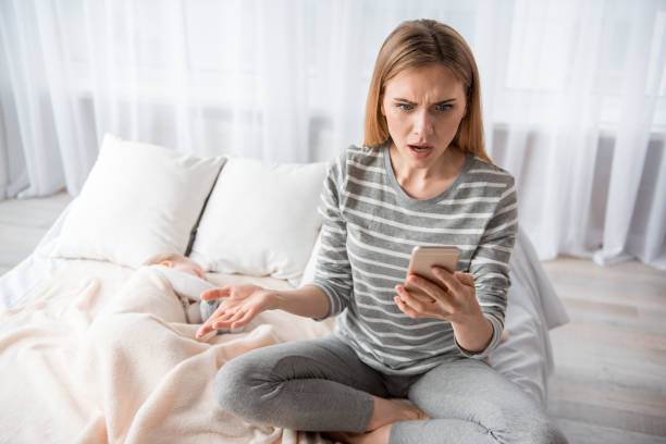 Astonished female using mobile phone Portrait of young woman sitting on sheets with smartphone in hand. She is looking at screen with surprised face. Baby is sleeping tremont stock pictures, royalty-free photos & images